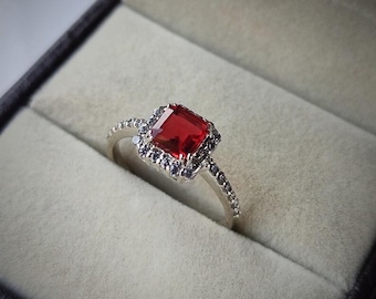 May include: A silver ring with a large, square-cut, red gemstone as the centerpiece. The gemstone is surrounded by smaller, dark-colored stones, and the band is adorned with tiny, clear stones. The ring is displayed in a black jewelry box.