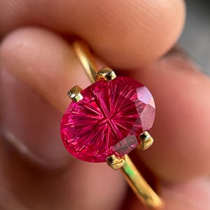 Peut inclure: Une bague en or orn&eacute;e d'une pierre pr&eacute;cieuse rose vif de forme ovale, dot&eacute;e d'un facettage en &eacute;toile ou en rayons de soleil. La pierre est maintenue par quatre petites griffes en or sur un anneau fin et poli, offrant une surface tr&egrave;s brillante.