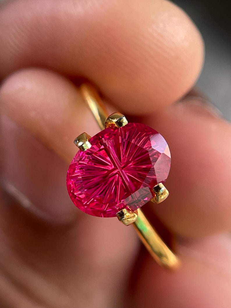 Peut inclure: Une bague en or orn&eacute;e d'une pierre pr&eacute;cieuse rose vif de forme ovale, dot&eacute;e d'un facettage en &eacute;toile ou en rayons de soleil. La pierre est maintenue par quatre petites griffes en or sur un anneau fin et poli, offrant une surface tr&egrave;s brillante.
