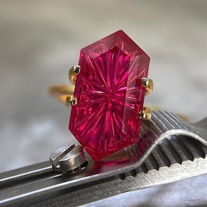 Puede incluir: Un anillo con una gema hexagonal alargada de color rosa intenso y facetas radiales intrincadas. La gema está asegurada en un engaste de metal dorado con cuatro garras, mostrando una superficie brillante que refleja la luz.