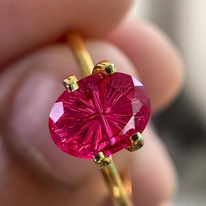 Peut inclure: Une bague en or orn&eacute;e d'une pierre pr&eacute;cieuse fuchsia de forme ovale, pr&eacute;sentant un motif de facettes en &eacute;toile ou en rayons de soleil. La pierre est maintenue par quatre petites griffes en or, sur un anneau en fil d'or poli et simple.