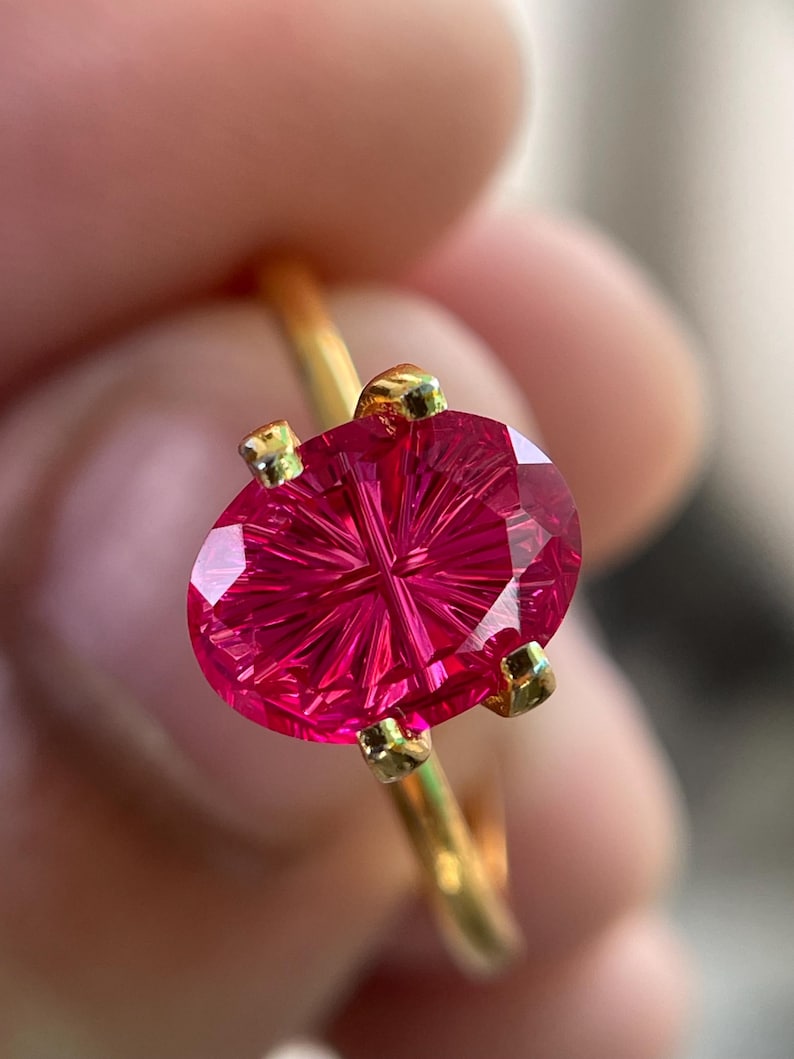 Peut inclure: Une bague en or orn&eacute;e d'une pierre pr&eacute;cieuse fuchsia de forme ovale, pr&eacute;sentant un motif de facettes en &eacute;toile ou en rayons de soleil. La pierre est maintenue par quatre petites griffes en or, sur un anneau en fil d'or poli et simple.