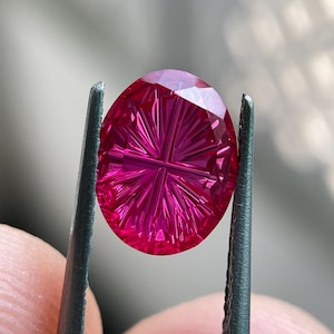 Peut inclure: Une pierre pr&eacute;cieuse ovale d'un rouge magenta vibrant, pr&eacute;sentant un motif de taille complexe en forme d'&eacute;toile sur sa surface. La pierre affiche une saturation profonde et une grande clart&eacute;, la lumi&egrave;re se refl&eacute;tant sur les facettes.