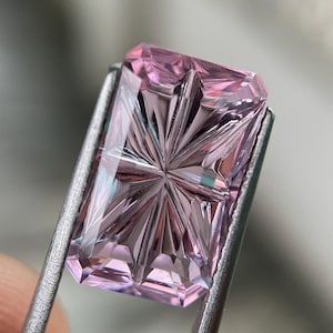 May include: A rectangular, light pink gemstone featuring a complex, star-like faceted pattern on its surface. The stone is held by metal tweezers, showcasing its translucent quality and sharp, geometric cuts that create a radiating light effect.