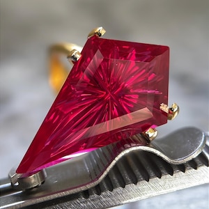 May include: A ring featuring a large, kite-shaped, vibrant red gemstone with a complex, star-like faceted cut. The gemstone is secured in a yellow gold setting with four prongs, showcasing a brilliant, reflective surface and sharp, geometric edges.