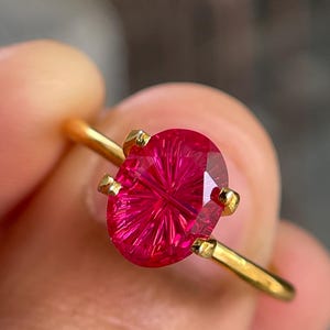 Op de afbeelding: Een gouden ring met een levendige, ovaal geslepen fuchsia edelsteen met een opvallend stervormig facetpatroon. De steen is gezet in een zetting met vier pootjes op een dunne, gepolijste gouden ring die een helder oppervlak vertoont.