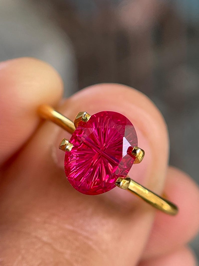 Peut inclure: Une bague en or orn&eacute;e d'une pierre pr&eacute;cieuse fuchsia de taille ovale avec un motif &agrave; facettes en forme d'&eacute;toile ou de rayons de soleil. La pierre est fix&eacute;e par quatre griffes sur un anneau en or fin et poli, offrant un &eacute;clat lumineux.