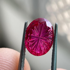 Peut inclure: Une pierre pr&eacute;cieuse ovale d'un rouge magenta vif pr&eacute;sentant un motif de facettes d&eacute;taill&eacute; en forme d'&eacute;toile ou de rayon de soleil sur sa surface. La pierre est tenue par des pinces m&eacute;talliques, r&eacute;v&eacute;lant sa translucidit&eacute; et ses coupes g&eacute;om&eacute;triques pr&eacute;cises.
