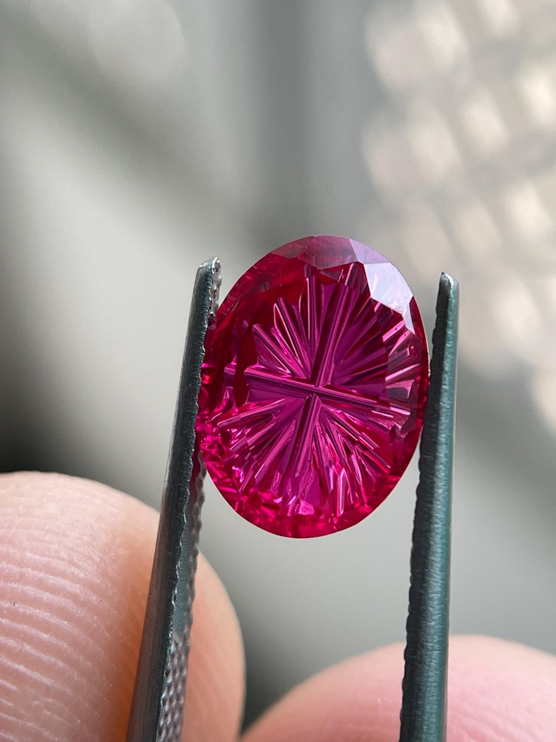 Peut inclure: Une pierre pr&eacute;cieuse ovale d'un rouge magenta vif pr&eacute;sentant un motif de facettes d&eacute;taill&eacute; en forme d'&eacute;toile ou de rayon de soleil sur sa surface. La pierre est tenue par des pinces m&eacute;talliques, r&eacute;v&eacute;lant sa translucidit&eacute; et ses coupes g&eacute;om&eacute;triques pr&eacute;cises.
