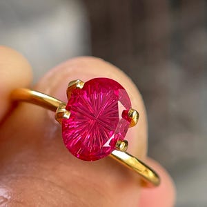 Peut inclure: Une bague en or orn&eacute;e d'une pierre pr&eacute;cieuse rose vif de forme ovale, pr&eacute;sentant un motif facett&eacute; radial en forme d'&eacute;toile. La pierre est maintenue par quatre petites griffes en or sur un anneau fin et poli, offrant un aspect brillant.