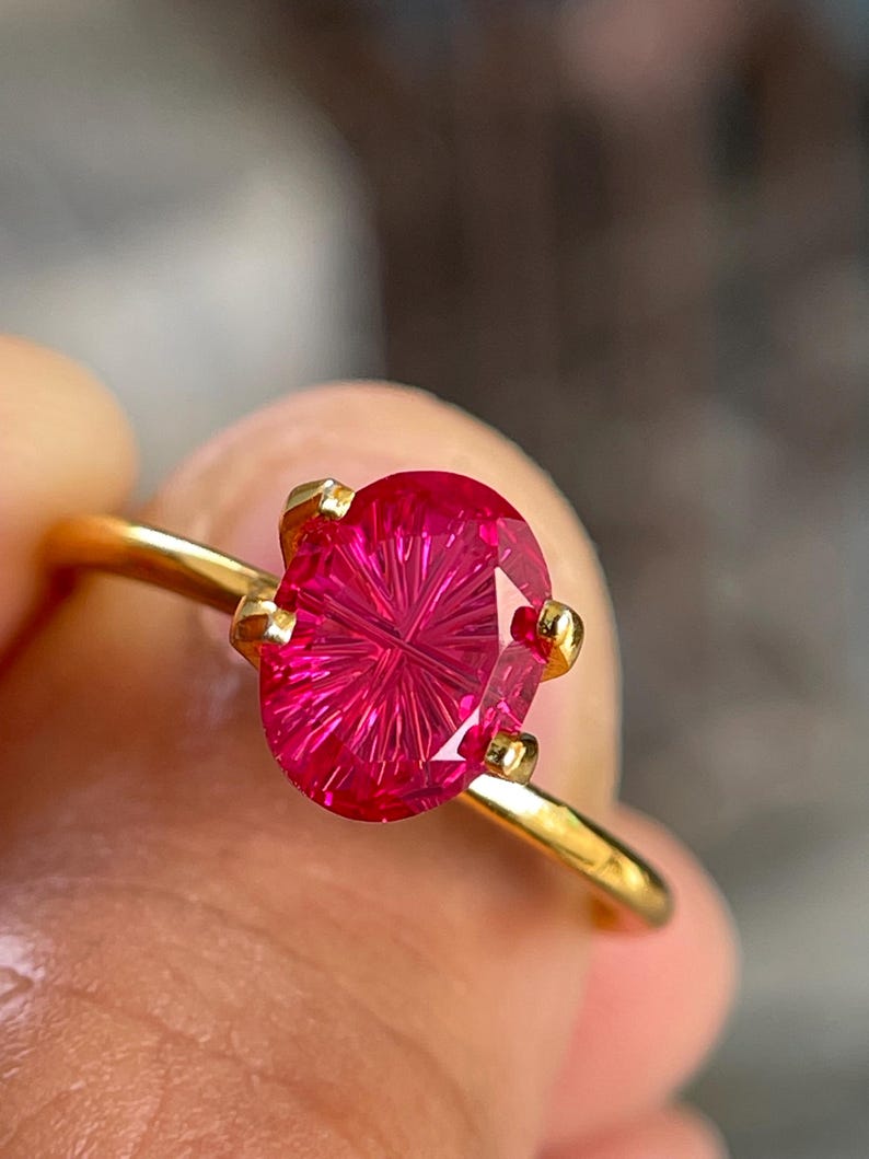 Peut inclure: Une bague en or orn&eacute;e d'une pierre pr&eacute;cieuse rose vif de forme ovale, pr&eacute;sentant un motif facett&eacute; radial en forme d'&eacute;toile. La pierre est maintenue par quatre petites griffes en or sur un anneau fin et poli, offrant un aspect brillant.
