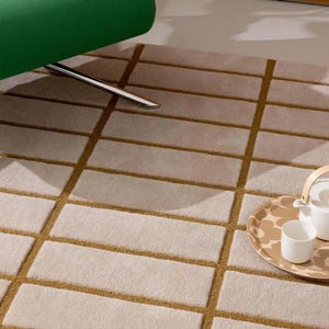 May include: A beige rectangular rug with a grid pattern of light brown lines. A green chair leg is visible in the upper left corner. A tray with a teapot and cups sits on the rug.