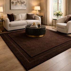 May include: A living room featuring a large, dark brown rectangular rug with a bordered design. The room includes a white sofa, two armchairs, a round coffee table, and a painting on the wall. The room is decorated in neutral tones.