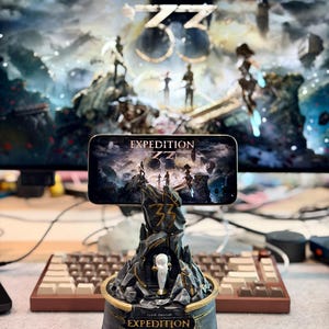 Puede incluir: Un soporte para teléfono Expedition 33 gris y dorado detallado. El soporte presenta un diseño de montaña rocosa con el texto "Expedition 33" y "Clair Obscur". Un teléfono muestra el mismo gráfico. Un monitor de computadora con el mismo gráfico está en el fondo.