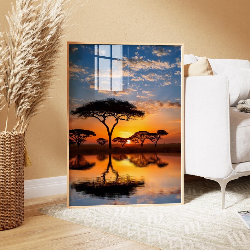 May include: Framed art print featuring a vibrant sunset over a tranquil lake. Silhouetted trees and their reflections are set against a sky of orange, yellow, and blue hues. The artwork is displayed in a light-colored wooden frame.