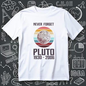 May include: White t-shirt featuring a retro design with the text "NEVER FORGET PLUTO 1930-2006". The design includes a planet graphic with a striped background in shades of orange, red, and blue. The shirt has a crew neck.