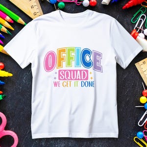Op de afbeelding: Wit t-shirt met de tekst "OFFICE SQUAD WE GET IT DONE" in kleurrijke letters. Het ontwerp gebruikt roze, oranje, groen, blauw en paars. Omringd door schoolbenodigdheden.