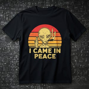 Puede incluir: Camiseta negra con un diseño retro. Presenta un extraterrestre fumando un cigarro y mostrando el dedo medio, sobre un fondo de puesta de sol. El texto "I CAME IN PEACE" está debajo.