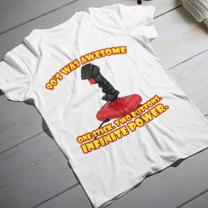 Può includere: T-shirt bianca con un joystick rosso e nero e la scritta "90's WAS AWESOME. ONE STICK. TWO BUTTONS. INFINITE POWER." Il testo e il joystick sono rossi e gialli.
