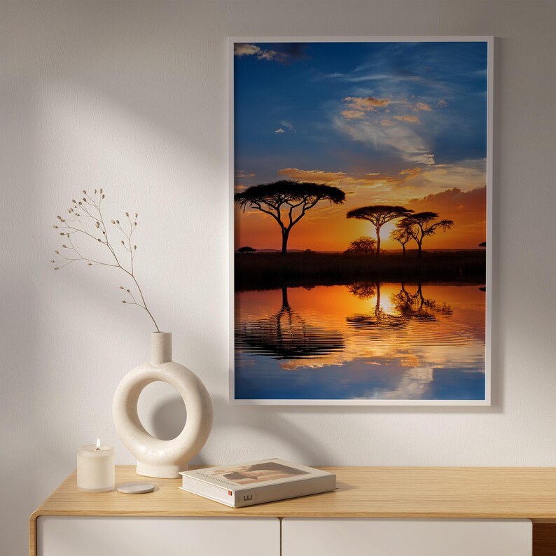 May include: A framed landscape print depicts a vibrant sunset over a body of water, reflecting the sky's orange and blue hues. Silhouetted trees stand on the horizon. A white vase, candle, and book sit on a wooden surface.