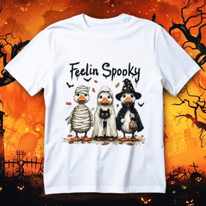Könnte beinhalten: Weißes T-Shirt mit einem Halloween-Motiv. Das Design zeigt drei Enten in Halloween-Kostümen: eine Mumie, ein Geist und eine Hexe, mit dem Text "Feelin Spooky". Die Enten stehen auf Herbstblättern.
