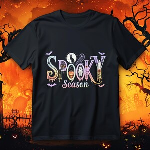 May include: Black t-shirt featuring the phrase "Spooky Season" in a decorative, multi-coloured font. The design incorporates a black cat, raven, ghost, and pumpkin. Bats and dripping paint elements enhance the Halloween aesthetic.