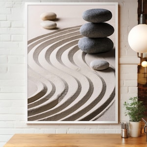 Könnte beinhalten: Ein gerahmter Kunstdruck eines Zen-Gartens. Das Bild zeigt einen Sandgarten mit geschwungenen Mustern und gestapelten grauen Steinen. Mehrere glatte, hellfarbige Steine sind verstreut, was ein Gefühl von Ruhe erzeugt.