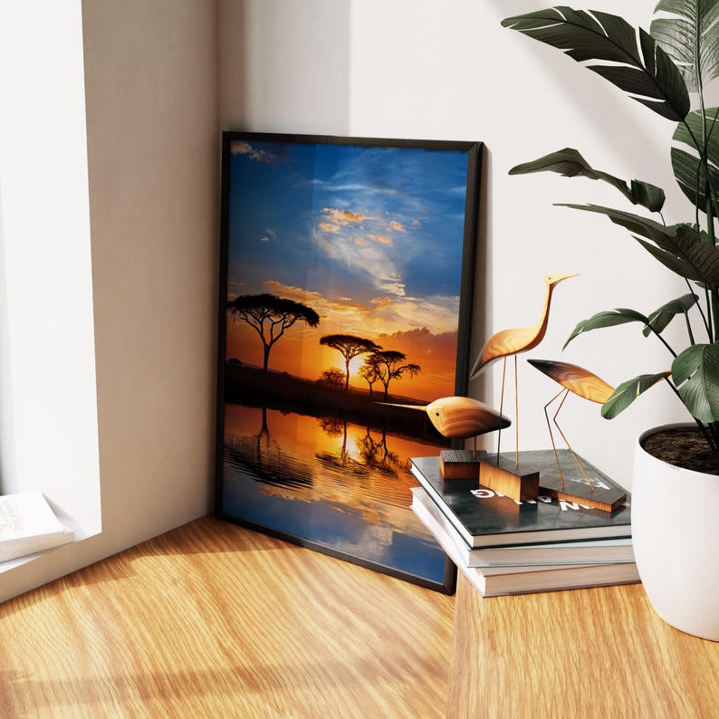 May include: A framed landscape print depicts a sunset over a body of water, with silhouetted trees. Wooden bird sculptures and a potted plant are nearby. The print is in a black frame and leans against a wall.