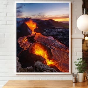 May include: A framed landscape photograph captures a volcanic eruption at sunset. Molten lava flows in fiery orange and red hues, contrasting with the dark, rocky terrain and a sky of blue, orange, and yellow.