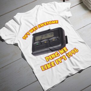 Może przedstawiać: Biała koszulka z czarną grafiką pagera oraz tekstem "90's WAS AWESOME" i "PING ME LIKE IT'S 1996" w kolorze żółtym i czerwonym. Pager wyświetla numery "8-35-49A".