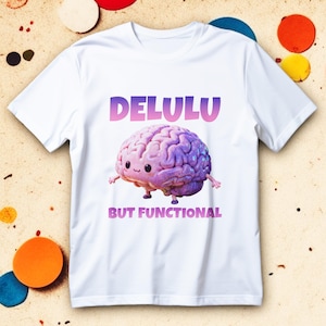 May include: White t-shirt featuring the text "DELULU" in a gradient purple and pink font above a cartoon brain with arms and legs. The phrase "BUT FUNCTIONAL" is below the brain in a similar font.