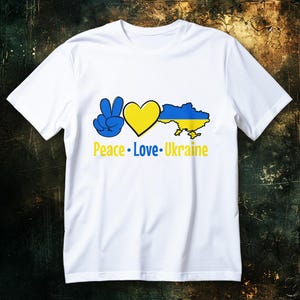 Puede incluir: Camiseta blanca con un signo de paz azul, un corazón amarillo y un mapa de Ucrania azul y amarillo. Las palabras "Peace Love Ukraine" están impresas en amarillo y azul. La camiseta es de material blanco suave.