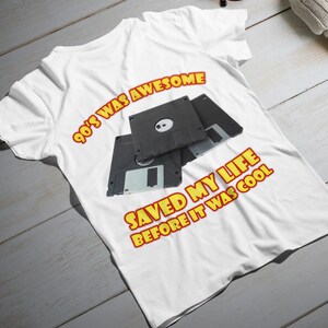 Może przedstawiać: Biała koszulka z grafiką trzech czarnych dyskietek. Koszulka ma tekst w kolorze czerwonym i żółtym z napisem "90's WAS AWESOME SAVED MY LIFE BEFORE IT WAS COOL."