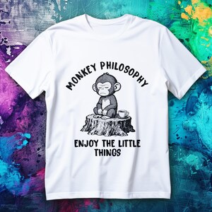 May include: White t-shirt featuring a black and white illustration of a meditating monkey on a tree stump, with the text "MONKEY PHILOSOPHY" above and "ENJOY THE LITTLE THINGS" below the image. A small cup of coffee is next to the monkey.