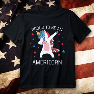Może przedstawiać: Czarny t-shirt z jednorożcem wykonującym dab, w patriotycznych kolorach, z napisem "PROUD TO BE AN AMERICORN". Jednorożec ma złoty róg, niebiesko-białe włosy oraz czerwone, białe i niebieskie akcenty. Czerwone i niebieskie gwiazdy otaczają jednorożca.