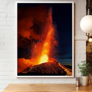 May include: A framed print of a volcanic eruption at night. The image captures the fiery orange and red lava spewing from the volcano against a dark blue sky. The eruption is the focal point, with glowing lava and smoke.