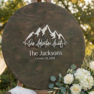 May include: A round wooden wedding sign with the text "Our Adventure Awaits" and "The Jacksons October 26, 2024". The sign features a mountain range design and is decorated with white flowers and greenery.