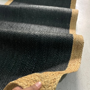 May include: A black jute runner rug with a natural tan border. The rug is made of woven jute fibers, creating a textured surface. The runner is folded over, showing the detail of the weave and the contrasting colors.