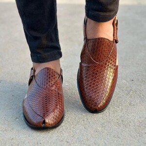 May include: A pair of brown leather Peshawari sandals. The sandals have a woven texture and a pointed toe. The shoes are worn by a person wearing black pants. The sandals have a strap with a gold buckle.
