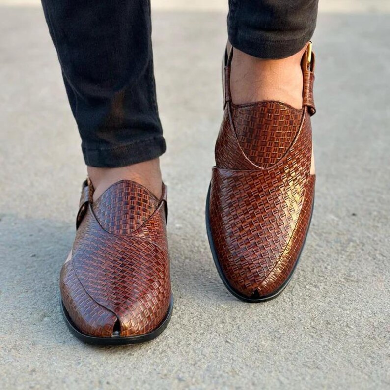 May include: A pair of brown leather Peshawari sandals. The sandals have a woven texture and a pointed toe. The shoes are worn by a person wearing black pants. The sandals have a strap with a gold buckle.