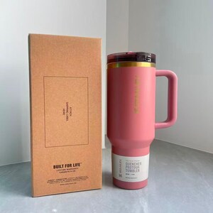 May include: A pink Stanley Quencher ProTour tumbler with a handle and a dark lid, positioned next to its rectangular cardboard packaging box. The box features the text BUILT FOR LIFE and ATTACH SHIPPING LABEL HERE. The tumbler holds 40 oz.