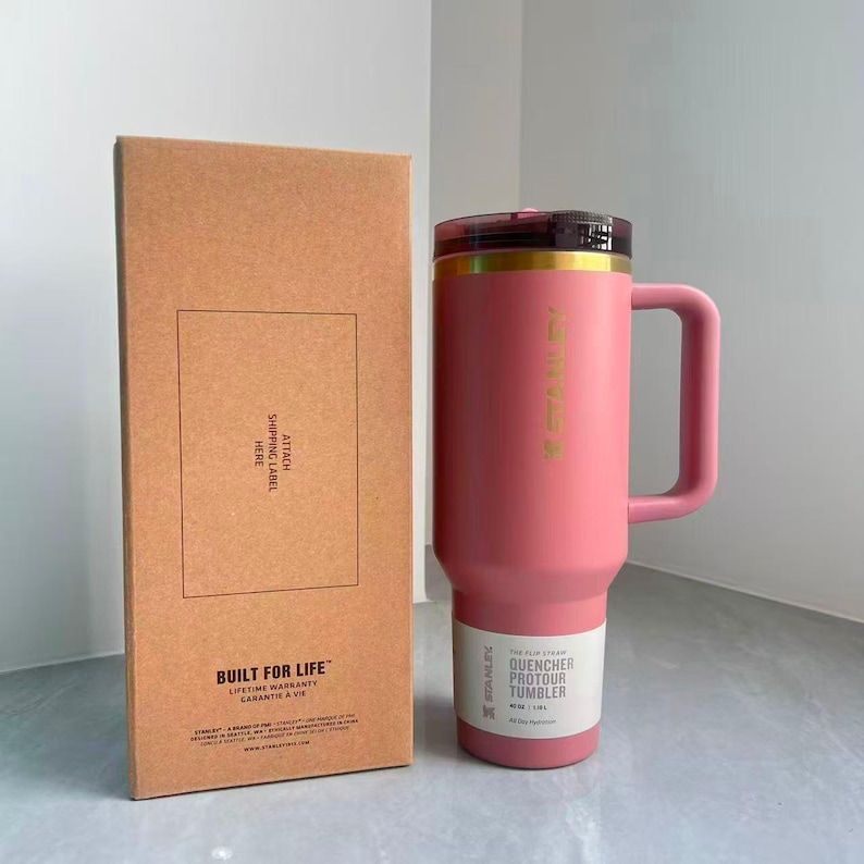May include: A pink Stanley Quencher ProTour tumbler with a handle and a dark lid, positioned next to its rectangular cardboard packaging box. The box features the text BUILT FOR LIFE and ATTACH SHIPPING LABEL HERE. The tumbler holds 40 oz.