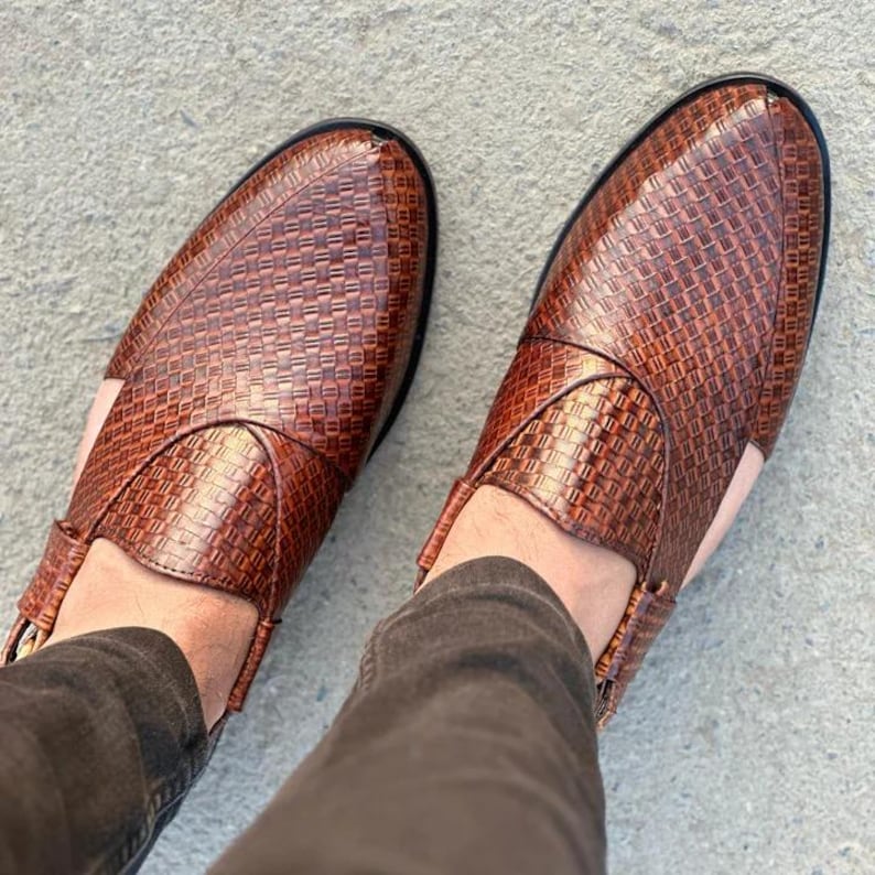 May include: A pair of brown leather slip-on shoes with a woven texture. The shoes have a rounded toe and a black sole. The shoes are worn by a person wearing brown pants.