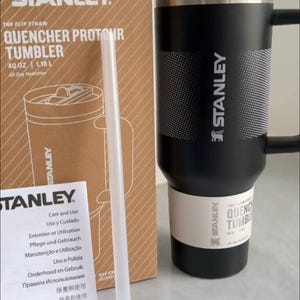 May include: A black Stanley Quencher ProTour tumbler with a handle, a black lid, and a white textured band. The tumbler is next to its cardboard packaging box, which displays the text Stanley Quencher ProTour 40 oz 1.18 L, and a clear straw.