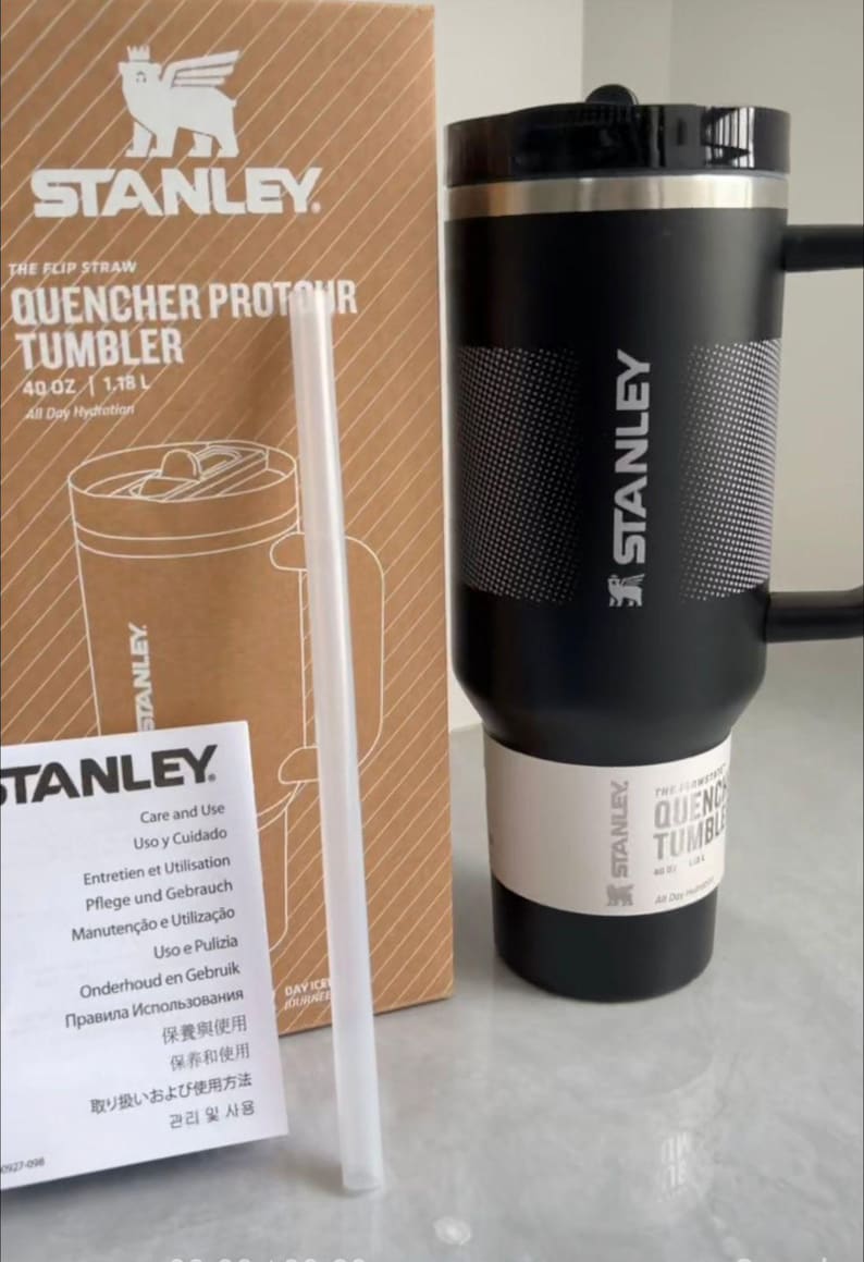 May include: A black Stanley Quencher ProTour tumbler with a handle, a black lid, and a white textured band. The tumbler is next to its cardboard packaging box, which displays the text Stanley Quencher ProTour 40 oz 1.18 L, and a clear straw.