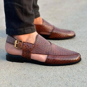 May include: Brown leather sandals with a woven pattern and a gold-colored buckle. The sandals have a black sole and a cutout design. The second sandal is slightly out of focus in the background.