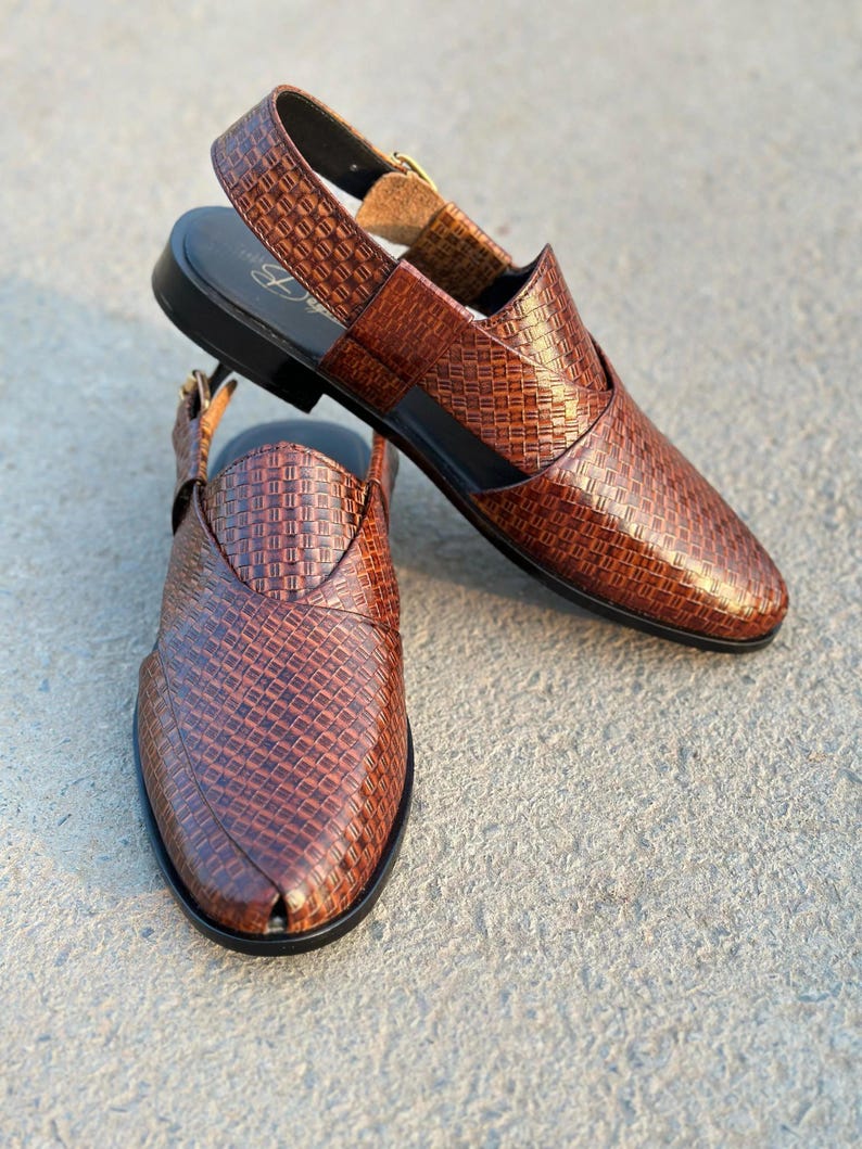 May include: Two brown leather sandals with a woven pattern. The sandals have a slingback strap with a buckle closure and a black sole. The shoes are displayed on a concrete surface.