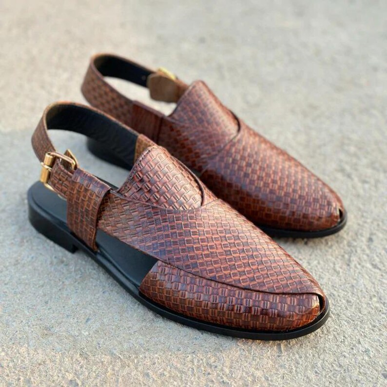 May include: A pair of brown leather sandals with a woven pattern. The sandals have a slingback strap with a gold-colored buckle and a black sole. The shoes are displayed on a gray surface.