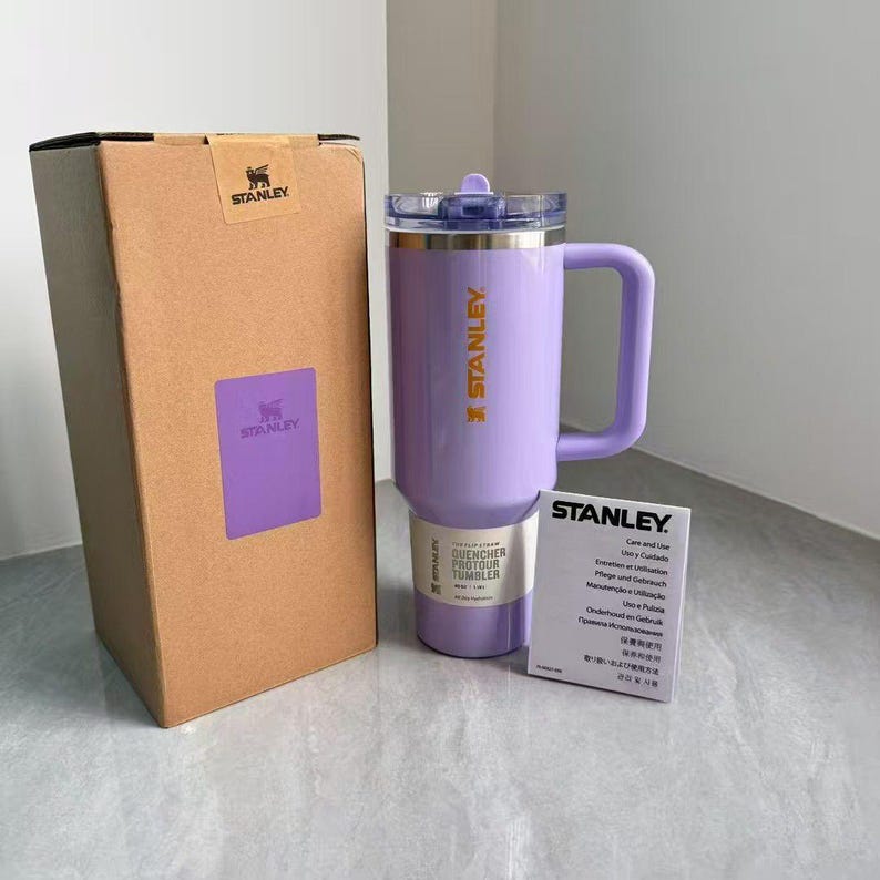 May include: A light purple Stanley Quencher H2.0 FlowState tumbler with a matching handle, a clear lid, and a straw. The tumbler is positioned next to its original brown cardboard packaging box and a white Stanley care and use instruction card.