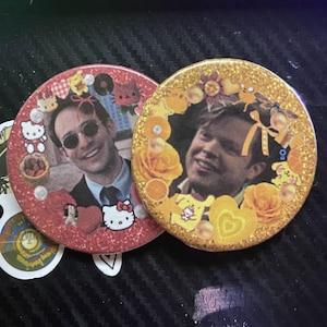 May include: Two round, glittery buttons. One is red with a photo of a man wearing sunglasses, surrounded by Hello Kitty and other cartoon characters. The other is gold with a photo of a man, surrounded by yellow roses and cartoon characters.
